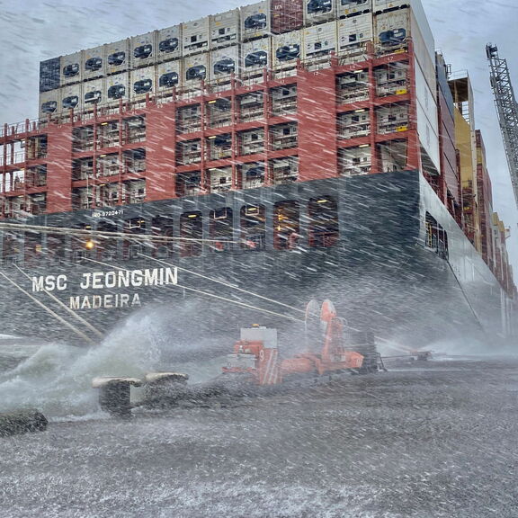 Containerschip MSC Jeongmin ligt aangemeerd aan de kade tijdens een zware storm, met opspattend water en oranje ShoreTension-units zichtbaar op de kade